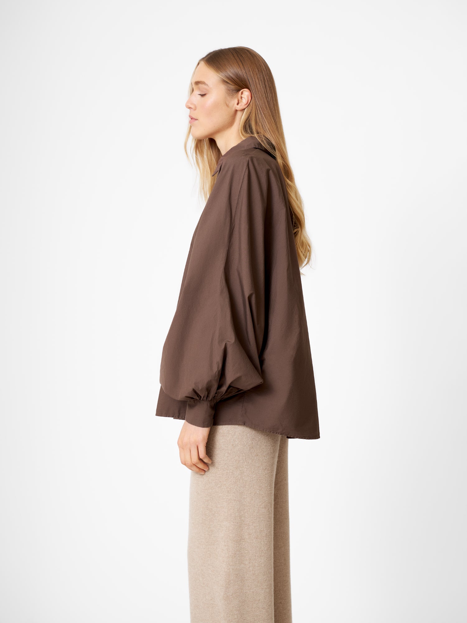 Eine Frau mit langen blonden Haaren steht im Profil vor einem unifarbenen Hintergrund und trägt die übergroße Bluse Antonia Espresso mit Fledermausärmeln und hellbeiger Hose.