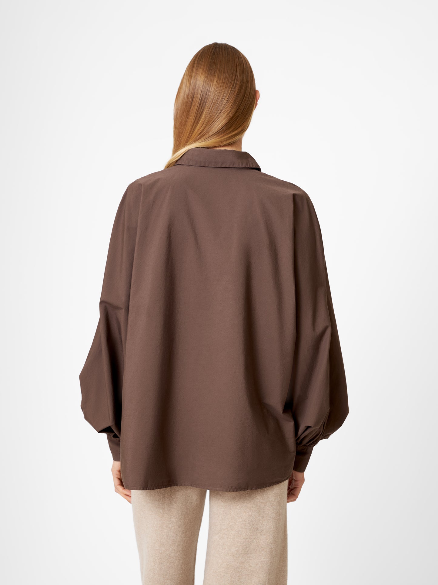 Eine Person mit langem, glattem hellbraunem Haar wird von hinten gezeigt. Sie trägt eine übergroße Bluse Antonia Espresso mit weiten Ärmeln und eine hellbeige Hose und steht vor einem schlichten weißen Hintergrund.