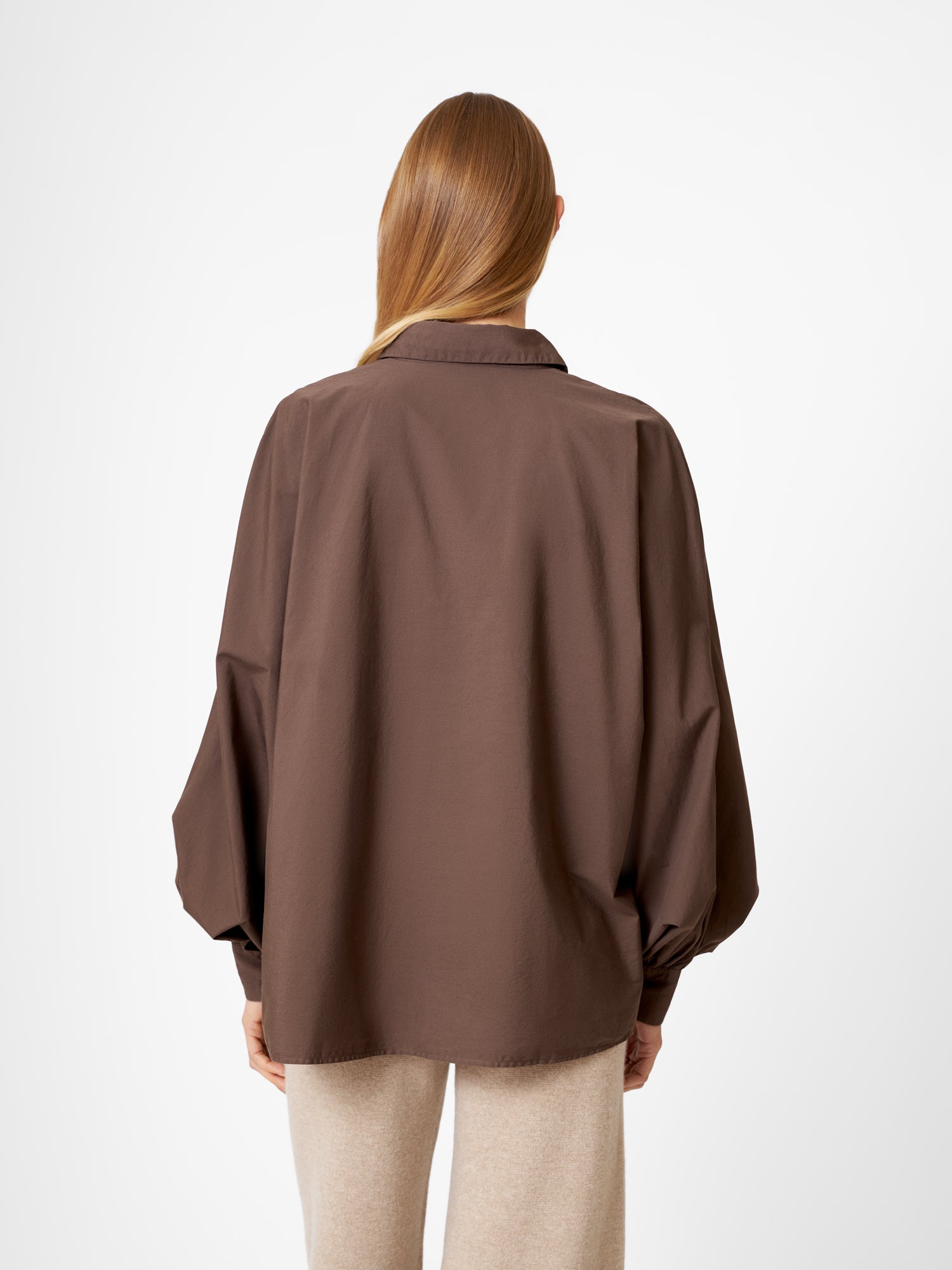 Eine Person mit langem, glattem hellbraunem Haar wird von hinten gezeigt. Sie trägt eine übergroße Bluse Antonia Espresso mit weiten Ärmeln und eine hellbeige Hose und steht vor einem schlichten weißen Hintergrund.