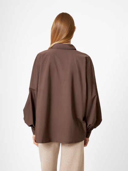 Eine Person mit langem, glattem hellbraunem Haar wird von hinten gezeigt. Sie trägt eine übergroße Bluse Antonia Espresso mit weiten Ärmeln und eine hellbeige Hose und steht vor einem schlichten weißen Hintergrund.