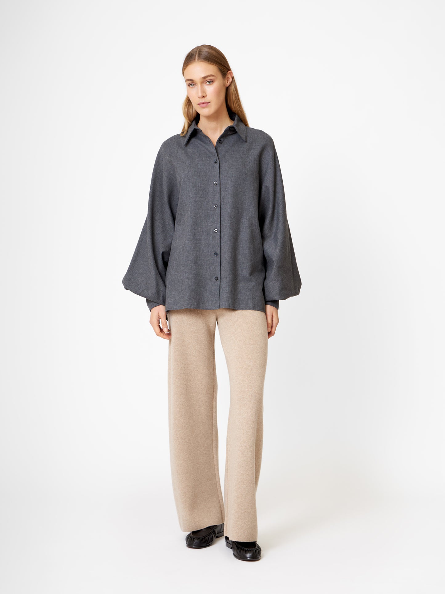 Eine Frau steht vor einem weißen Hintergrund und trägt die Bluse Antonia Flanell Anthra mit Ballonärmeln, eine beigefarbene Hose mit weitem Bein und schwarze Schuhe.