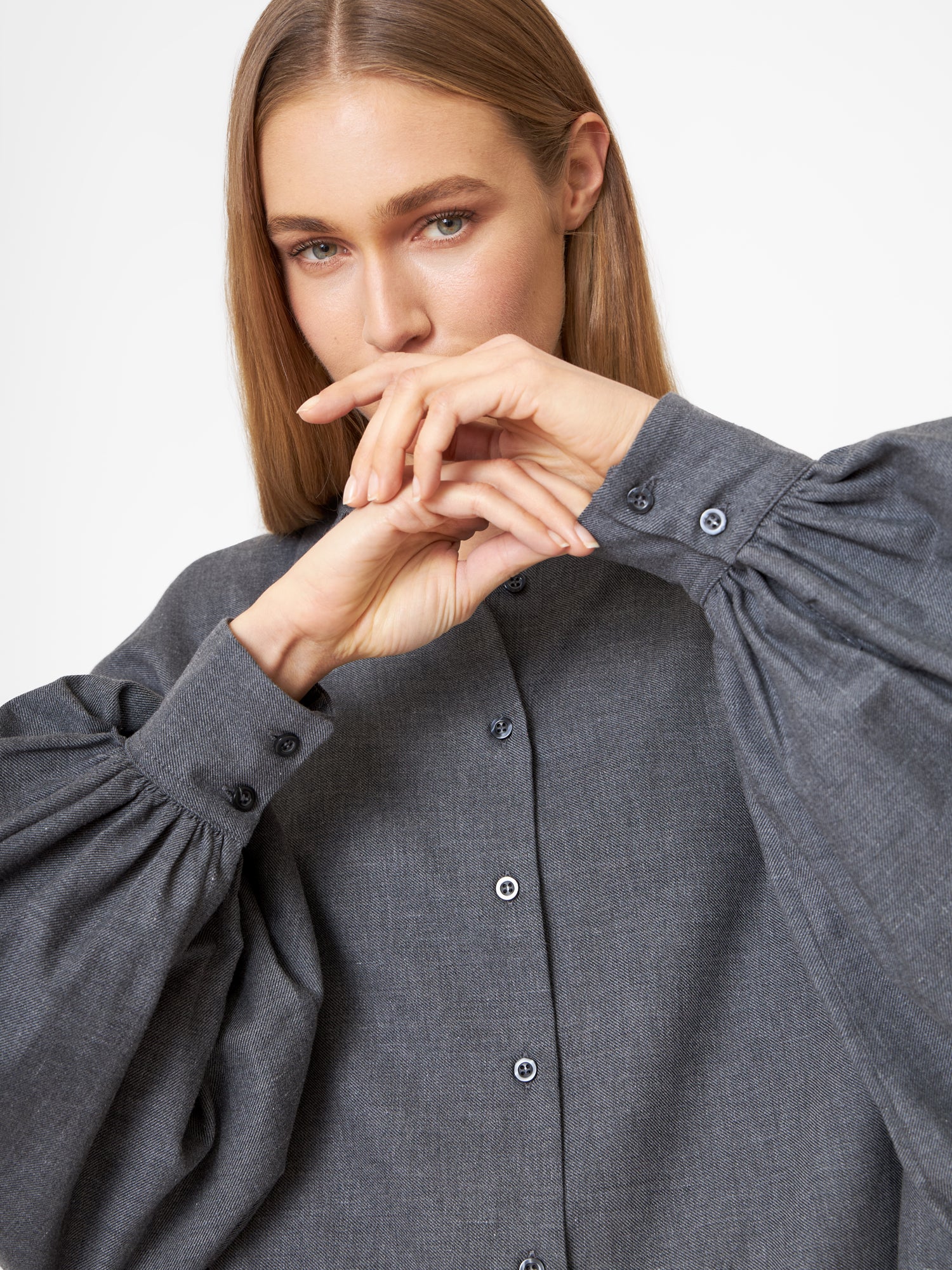 Eine Frau mit glattem, hellbraunem Haar trägt die Bluse Antonia Flanell Anthra mit voluminösen Ärmeln in zartem Grau und hält ihre Hände anmutig vor einem schlichten weißen Hintergrund neben ihrem Gesicht.