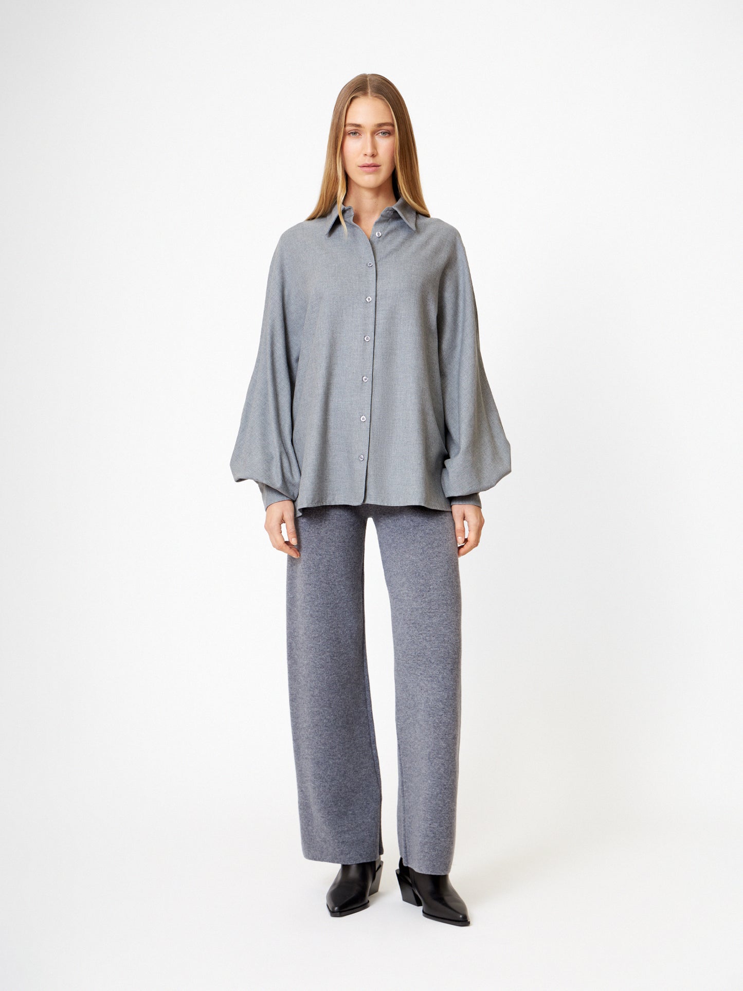 Eine Frau steht vor einem weißen Hintergrund und trägt die Bluse Antonia Grey Flanell, eine elegante graue Bluse, die mit einer grauen Hose mit weitem Bein und schwarzen Stiefeln kombiniert ist, und schaut mit neutralem Blick geradeaus.