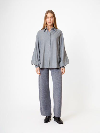 Eine Frau steht vor einem weißen Hintergrund und trägt die Bluse Antonia Grey Flanell, eine elegante graue Bluse, die mit einer grauen Hose mit weitem Bein und schwarzen Stiefeln kombiniert ist, und schaut mit neutralem Blick geradeaus.