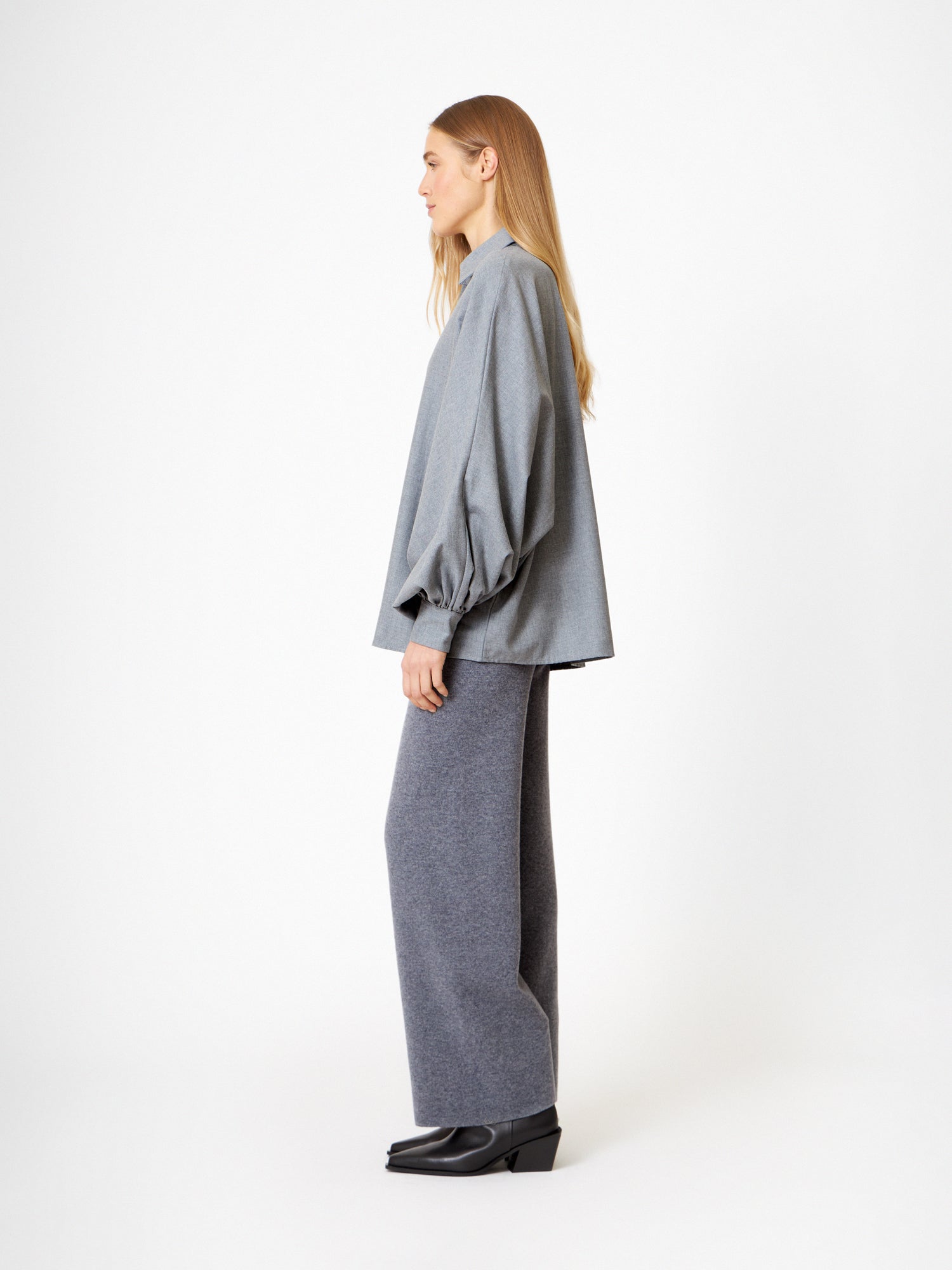 Eine Frau steht im Profil vor einem weißen Hintergrund und trägt einen Bluse Antonia Grey Flanell, eine graue Hose mit weitem Bein und schwarze Schuhe. Sie hat glattes, blondes Haar, das nach unten getragen wird.