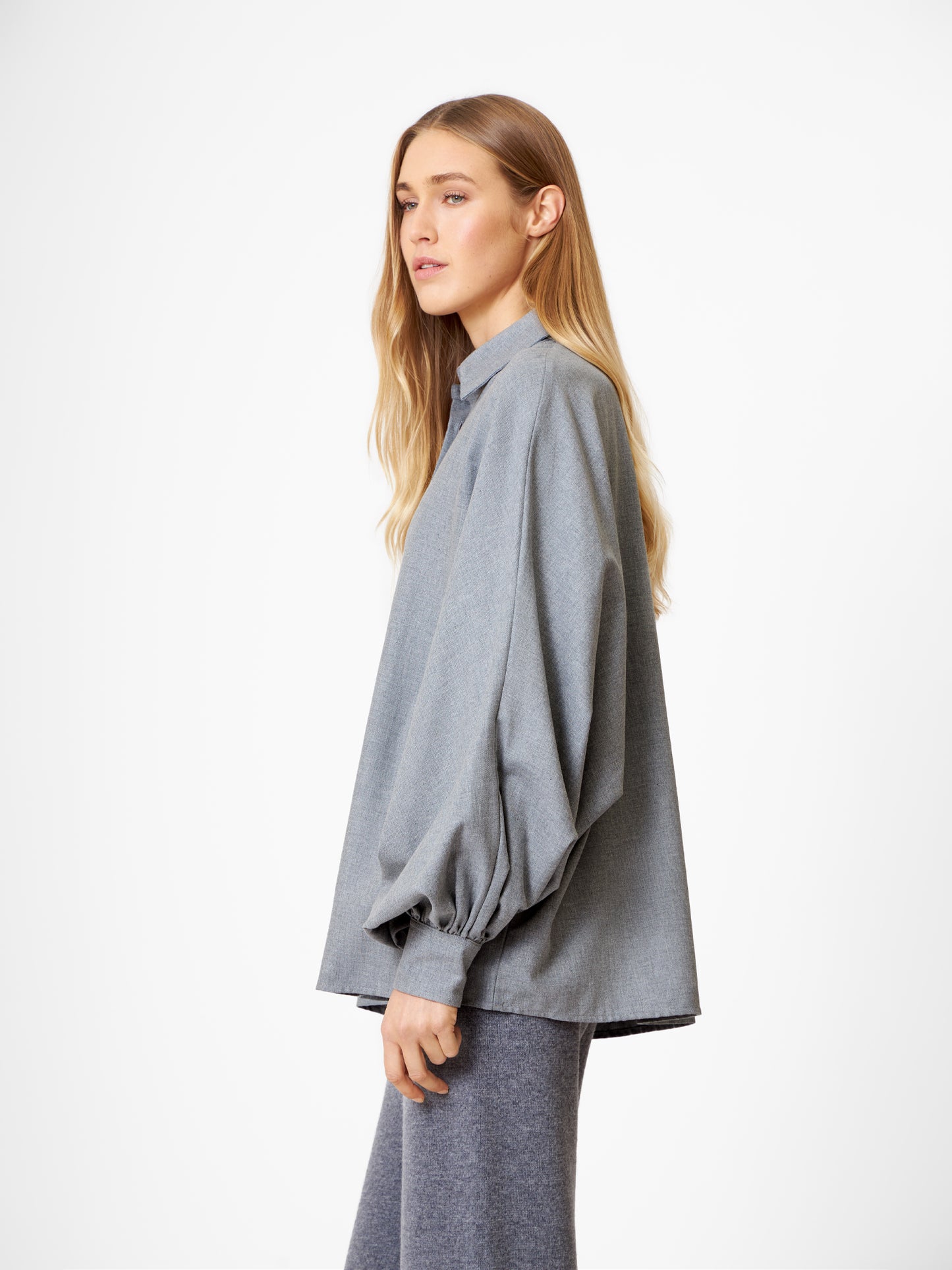 Eine Frau mit langem blondem Haar trägt die Bluse Antonia Grey Flanell mit voluminösen Ärmeln und passender grauer Hose, die im Profil vor einem schlichten weißen Hintergrund steht.