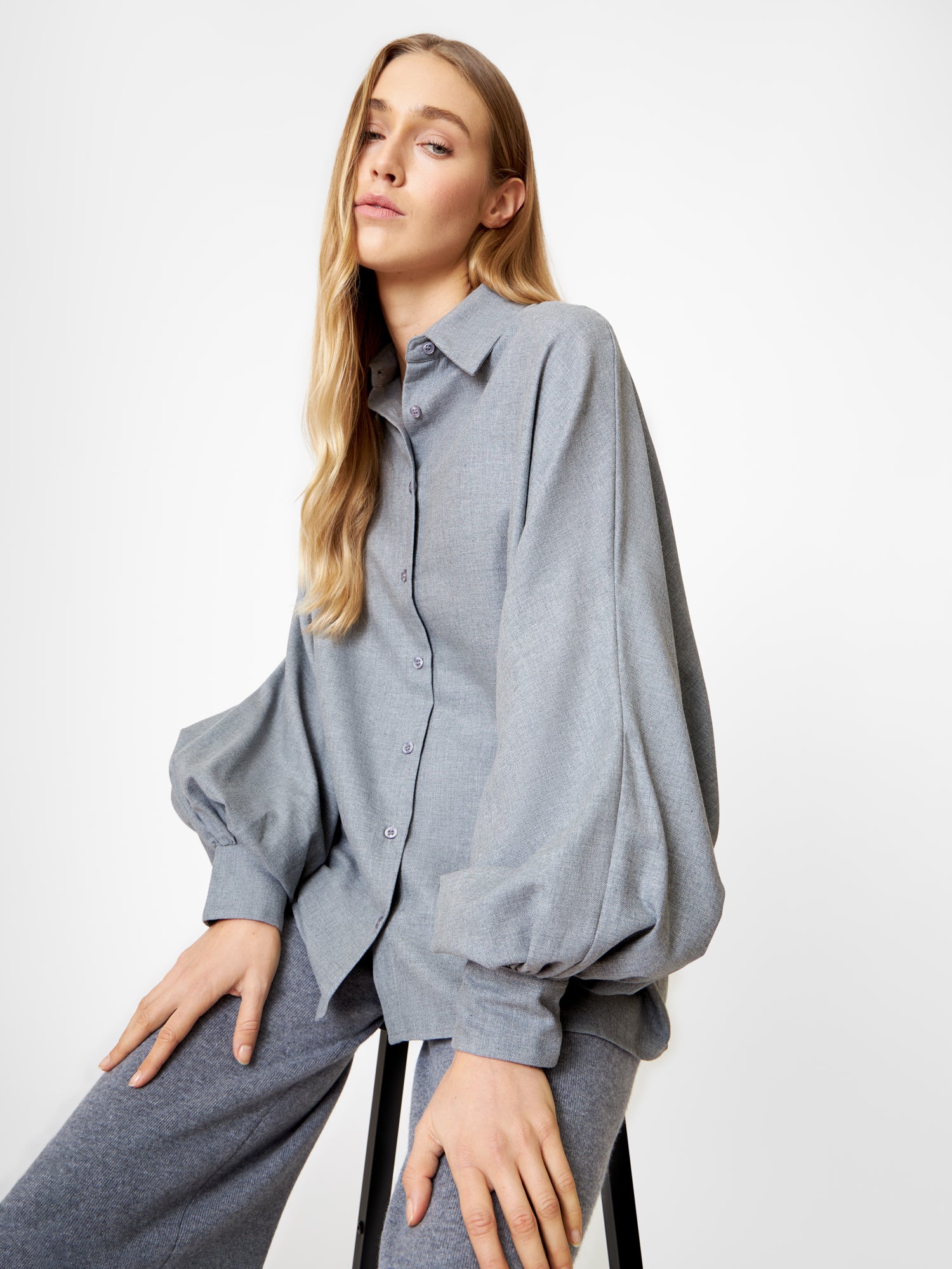 Eine Frau mit langen blonden Haaren sitzt auf einem Hocker und trägt die Bluse Antonia Grey Flanell - eine lockere, elegante graue Bluse mit weiten Ärmeln - und eine dazu passende Hose vor einem schlichten weißen Hintergrund.