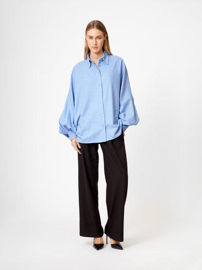 Eine Person steht vor einem schlichten Hintergrund und trägt die Bluse Antonia Flanell Light blue, kombiniert mit einer schwarzen Hose mit weitem Bein und schwarzen spitzen Absätzen.