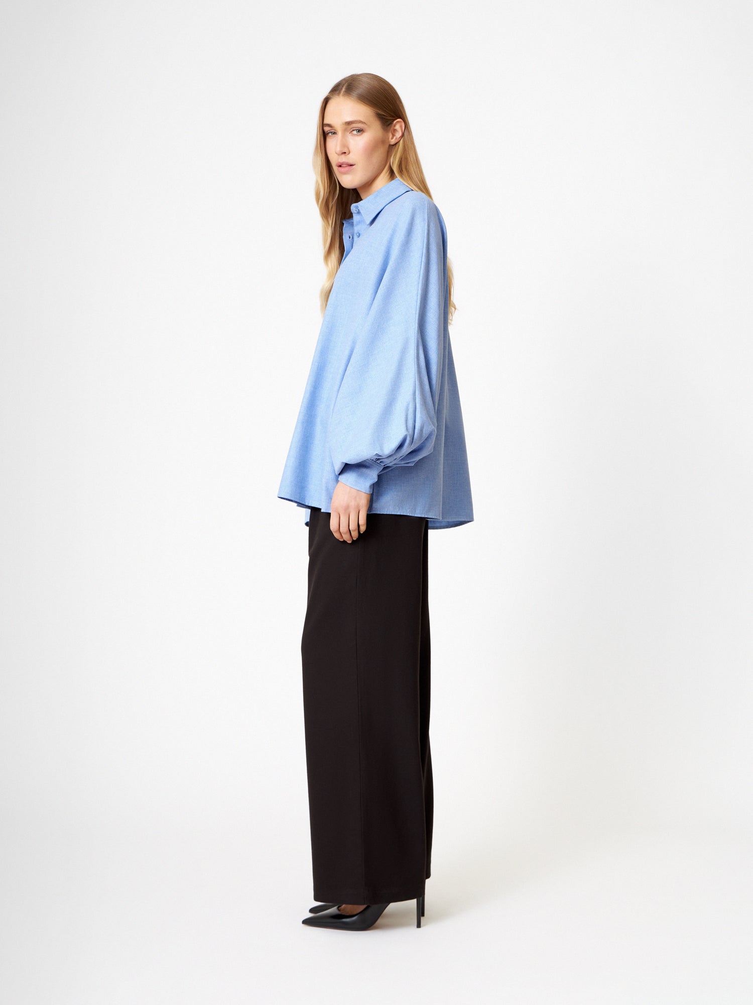Eine Frau steht seitlich vor einem schlichten weißen Hintergrund und trägt die Bluse Antonia Flanell Hellblau mit Ballonärmeln, gepaart mit einer schwarzen Hose mit weitem Bein und schwarzen Absätzen.