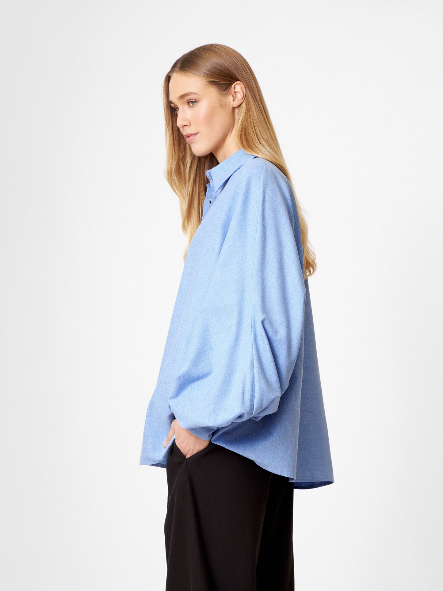 Eine Frau mit langen blonden Haaren modelliert die Bluse Antonia Flanell Hellblau, ein locker sitzendes Button-up-Hemd mit voluminösen Ärmeln, gepaart mit einer schwarzen Hose. Sie steht seitlich mit einer Hand in der Tasche vor einem weißen Hintergrund.