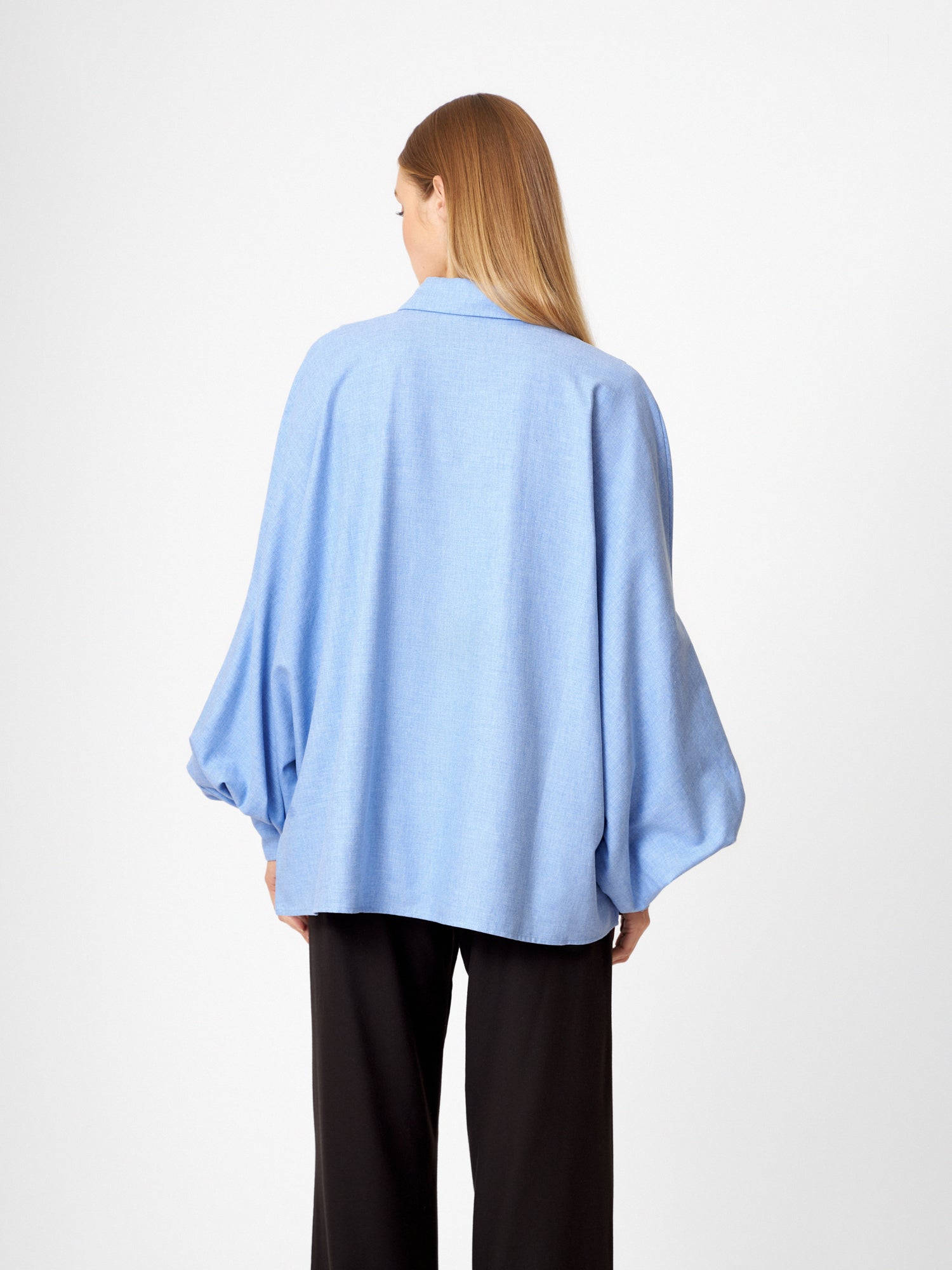 Eine Frau mit glattem, langem blondem Haar steht mit dem Rücken zur Kamera. Sie trägt die Bluse Antonia Flanell Light blue, gepaart mit einer schwarzen Hose, vor einem schlichten weißen Hintergrund.