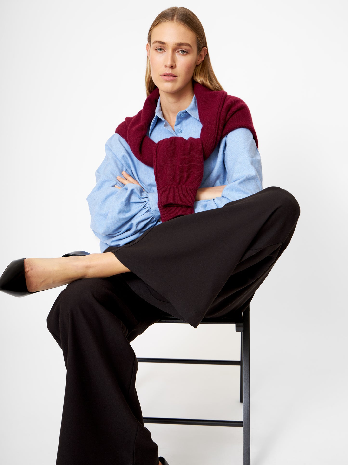 Eine Frau sitzt mit gekreuzten Beinen auf einem Stuhl und trägt die Bluse Antonia Flanell in Hellblau. Dazu trägt sie eine schwarze Hose mit weitem Bein, schwarze Absätze und einen burgunderroten Pullover über den Schultern und schaut mit neutralem Blick in die Kamera.