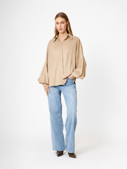 Eine Frau steht vor einem weißen Hintergrund und trägt ein übergroßes Bluse Antonia Vegan Leather Beige mit weiten Ärmeln, hellblaue Jeans und schwarze Schuhe mit spitzen Zehen. Ihr Haar ist glatt und sie posiert mit einer Hand in der Tasche für einen entspannten Look.