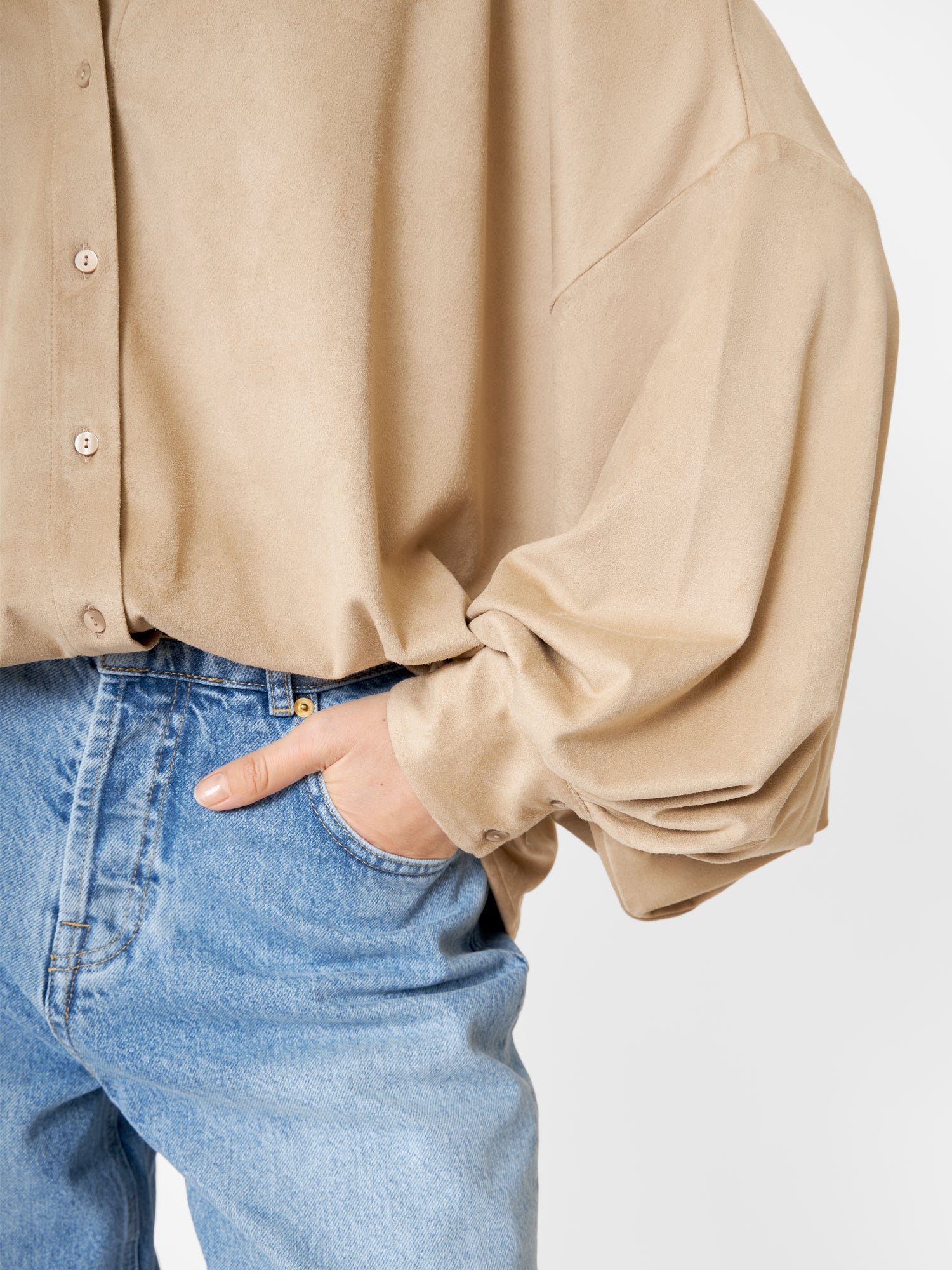 Eine Person trägt eine übergroße Bluse Antonia Vegan Leather Beige mit der linken Hand in der Tasche einer hellblauen Jeans und steht vor einem schlichten weißen Hintergrund.