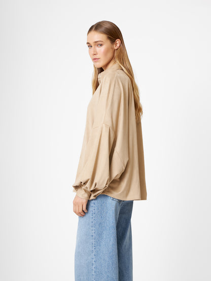 Eine Frau mit langem, hellbraunem Haar steht seitlich und trägt ein Bluse Antonia Vegan Leather Beige in einer lockeren, übergroßen Passform und hellblaue Jeans mit weitem Bein vor einem schlichten weißen Hintergrund.