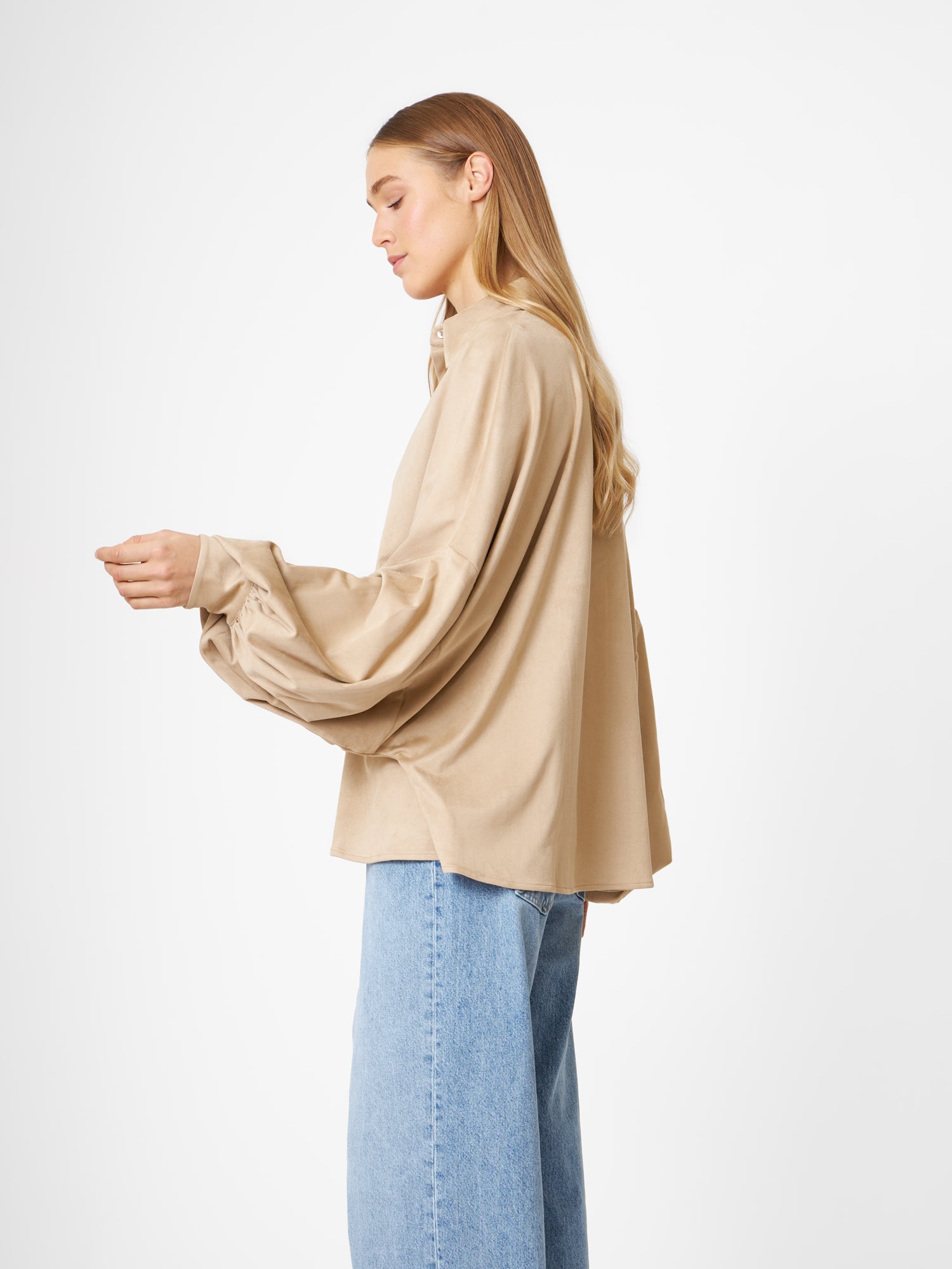 Eine Frau mit langen blonden Haaren trägt die Bluse Antonia Vegan Leather Beige in One Size mit hellblauen Jeans, steht vor einem schlichten weißen Hintergrund, blickt nach links und schaut nach unten.