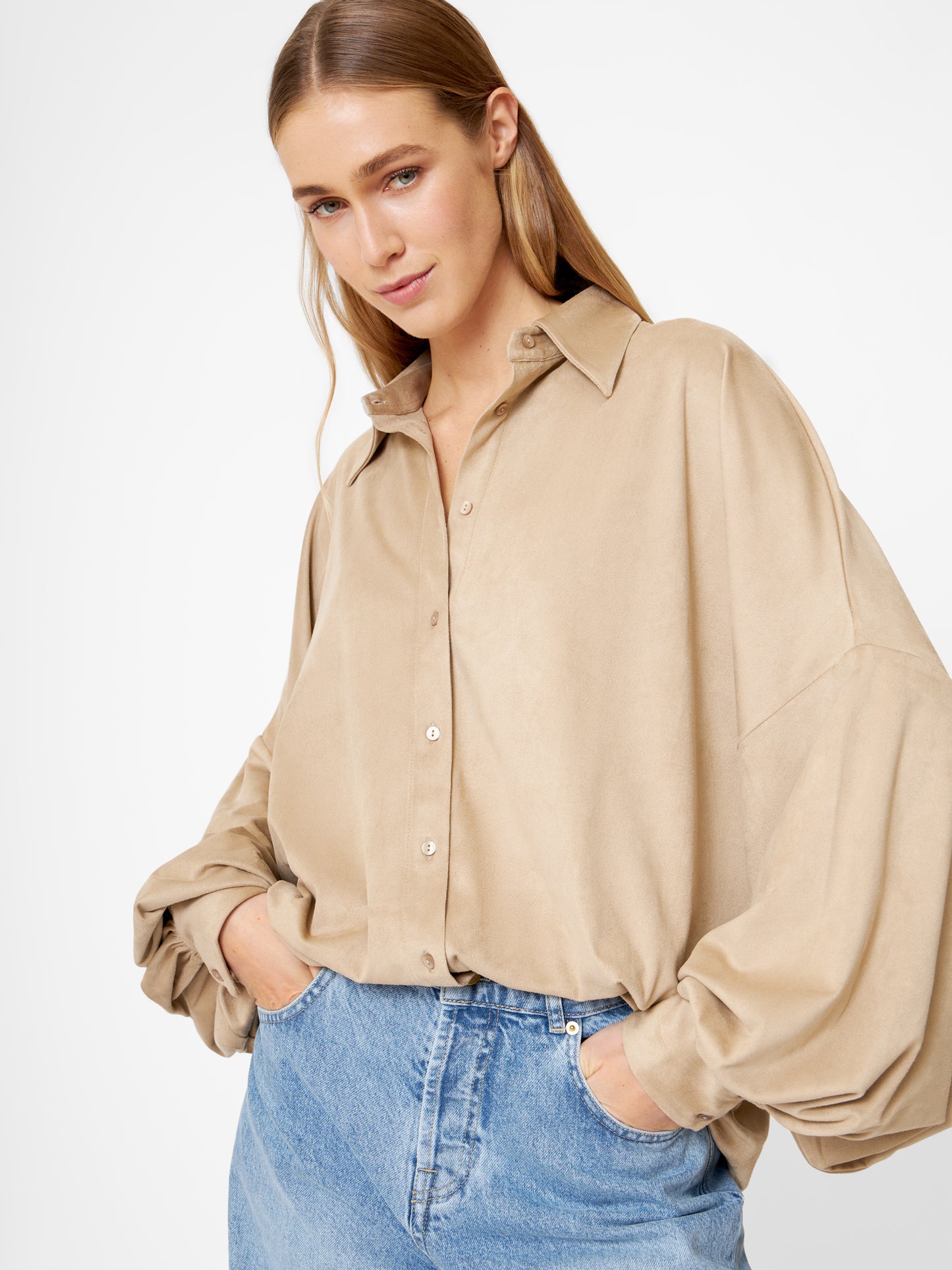 Eine Frau mit langem, glattem blondem Haar trägt eine übergroße Bluse Antonia Vegan Leather Beige mit Ballonärmeln, die in eine hellblaue Jeans gesteckt wird. Sie steht mit den Händen in den Taschen vor einem schlichten, hellen Hintergrund.