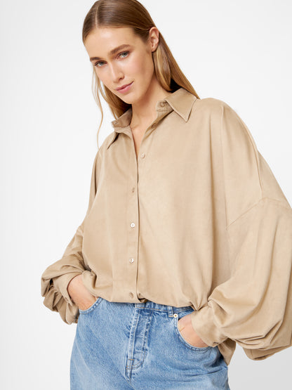 Eine Frau mit langem, glattem blondem Haar trägt eine übergroße Bluse Antonia Vegan Leather Beige mit Ballonärmeln, die in eine hellblaue Jeans gesteckt wird. Sie steht mit den Händen in den Taschen vor einem schlichten, hellen Hintergrund.