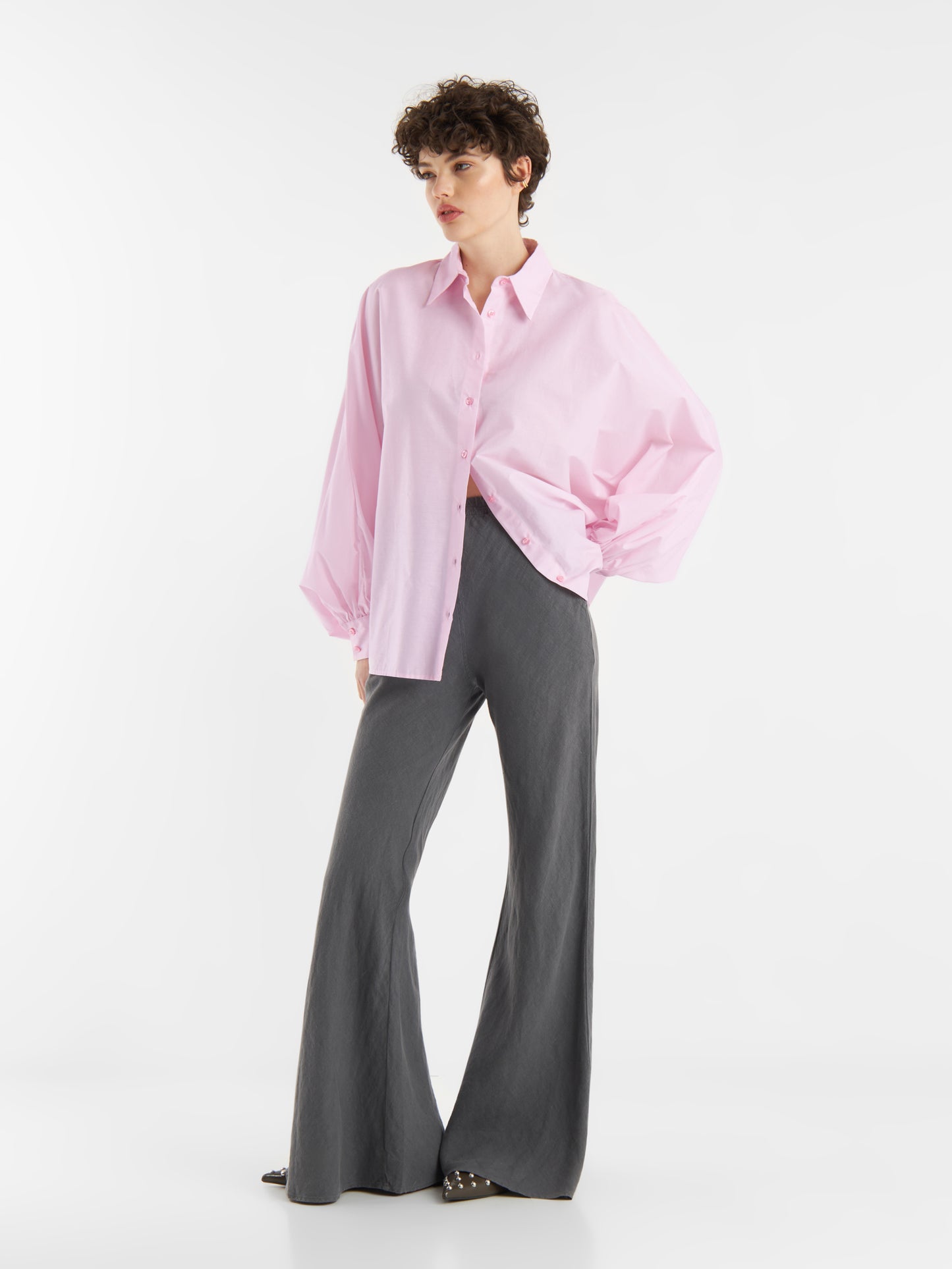 Eine Person, die die übergroße Bluse Antonia Rosa und eine dunkelgraue Hose mit weitem Bein trägt, steht vor einem schlichten weißen Hintergrund und schaut mit einer Hand in der Tasche zur Seite.