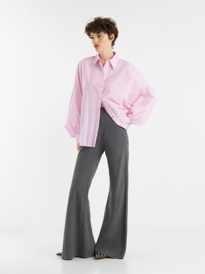 Eine Person, die die übergroße Bluse Antonia Rosa und eine dunkelgraue Hose mit weitem Bein trägt, steht vor einem schlichten weißen Hintergrund und schaut mit einer Hand in der Tasche zur Seite.