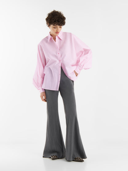 Eine Person mit kurzen lockigen Haaren trägt eine übergroße Bluse Antonia Rosa, eine graue Schlaghose und Nietenschuhe. Sie steht vor einem schlichten weißen Hintergrund und blickt nach unten.