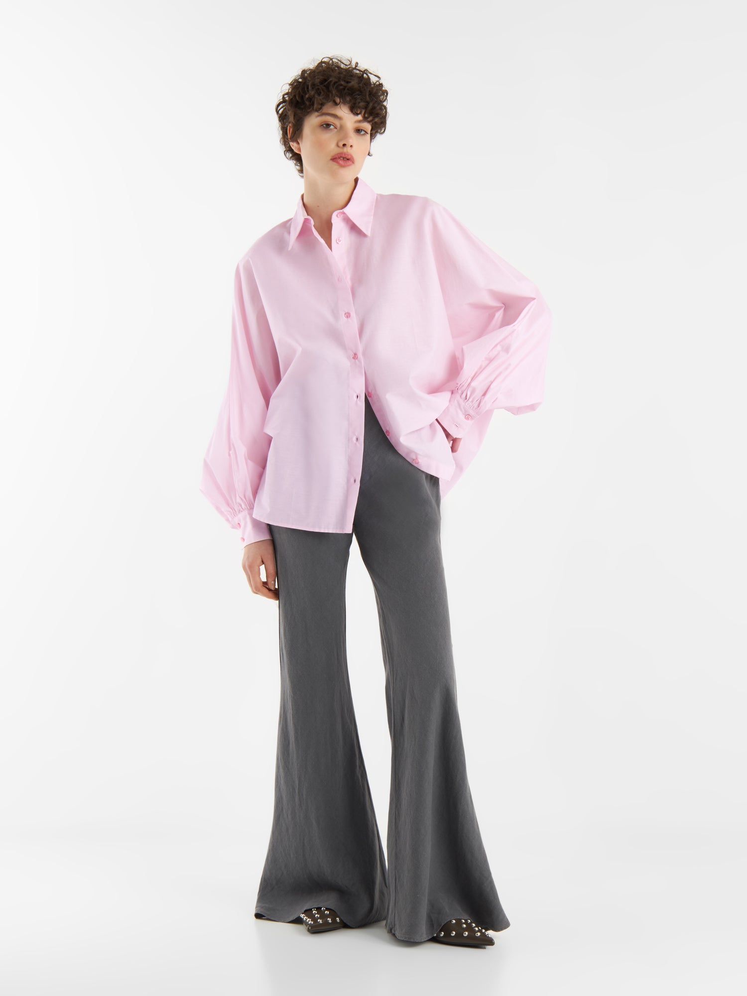 Eine Person steht vor einem weißen Hintergrund und trägt eine übergroße Bluse Antonia Rosa mit weiten Ärmeln, graue Schlaghosen und schwarze Nietenschuhe. Die entspannte Pose zeigt eine Hand in einer Tasche.