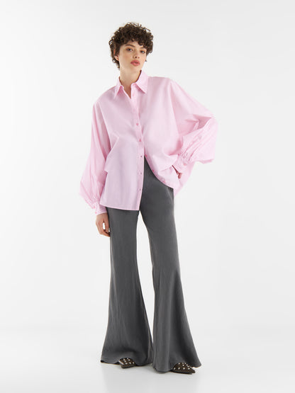 Eine Person steht vor einem weißen Hintergrund und trägt eine übergroße Bluse Antonia Rosa mit weiten Ärmeln, graue Schlaghosen und schwarze Nietenschuhe. Die entspannte Pose zeigt eine Hand in einer Tasche.