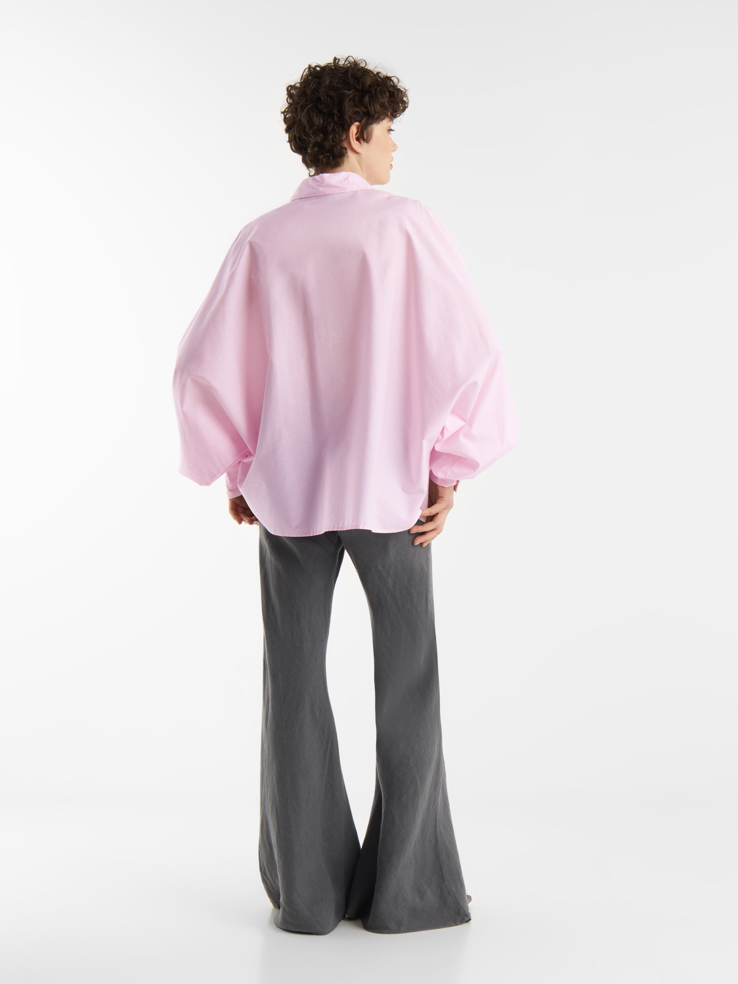 Eine Person mit kurzen lockigen Haaren steht mit dem Gesicht zur Wand und trägt eine übergroße Bluse Antonia Rosa mit weiten Ärmeln und grauen Schlaghosen vor einem schlichten weißen Hintergrund.