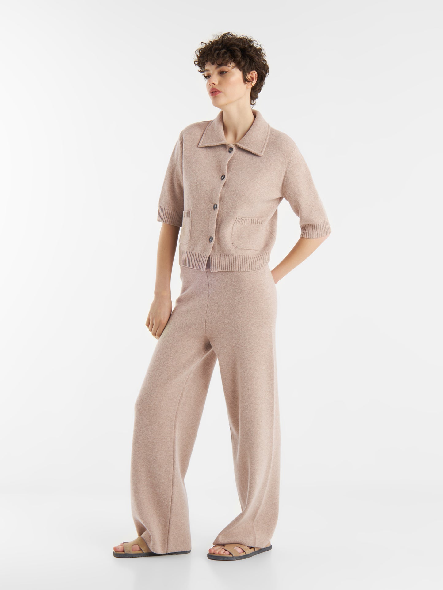 Eine Person mit kurzen lockigen Haaren trägt die beigefarbene Jacke Lilly und eine dazu passende Hose mit weitem Bein und Sandalen, die vor einem schlichten weißen Hintergrund stehen.