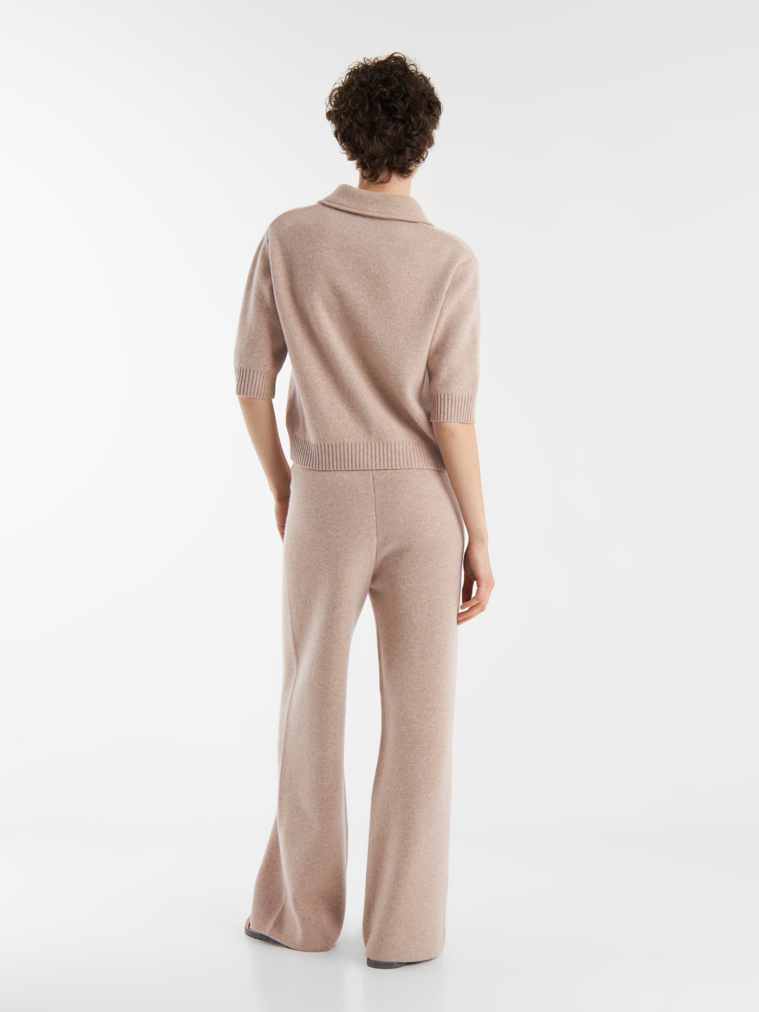 Eine Person mit kurzen lockigen Haaren steht mit dem Gesicht zur Wand und trägt einen hellbeigen, kurzärmeligen Strickpullover, eine passende Strickhose mit weitem Bein und die Jacke Lilly beige, die vor einem einfarbig weißen Hintergrund gestylt ist.