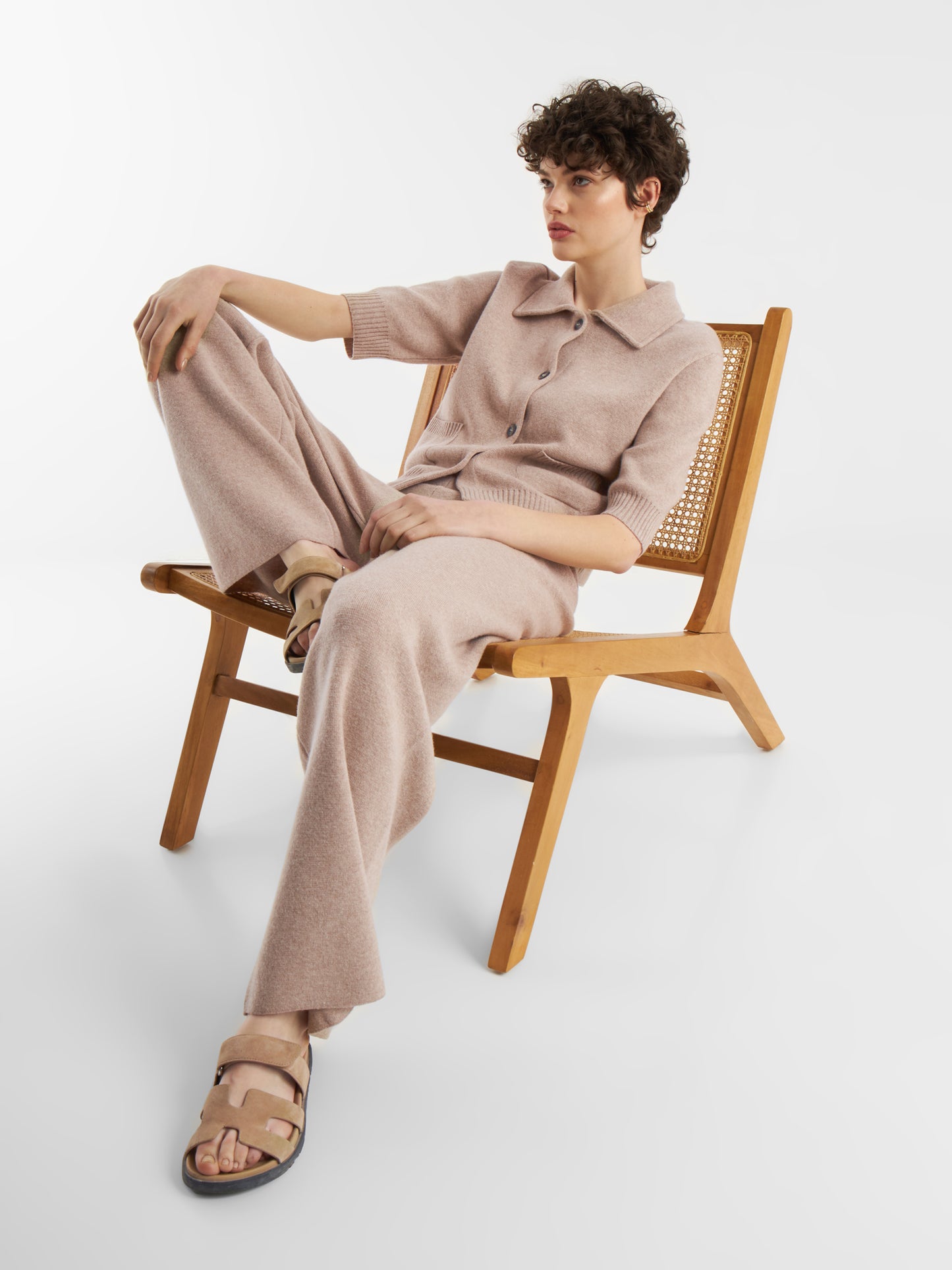 Eine Person mit kurzen lockigen Haaren, bekleidet mit einem beigefarbenen Strickoberteil und einer beigen Merino-Hose, sitzt entspannt auf einem Holzstuhl mit gekreuzten Beinen und trägt beigefarbene Sandalen vor einem schlichten weißen Hintergrund.