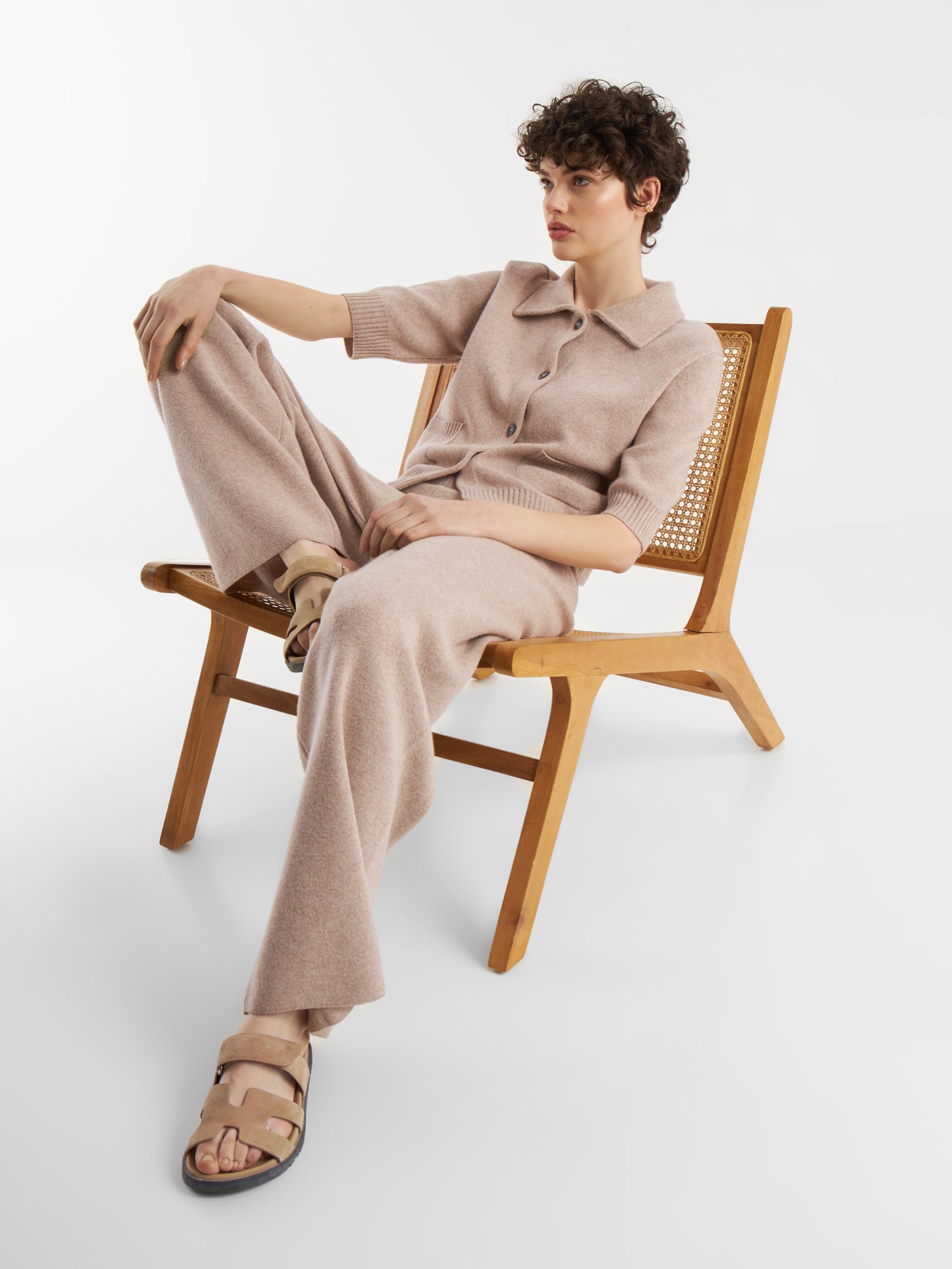 Eine Person mit kurzen lockigen Haaren, bekleidet mit einem beigefarbenen Strickoberteil und einer beigen Merino-Hose, sitzt entspannt auf einem Holzstuhl mit gekreuzten Beinen und trägt beigefarbene Sandalen vor einem schlichten weißen Hintergrund.