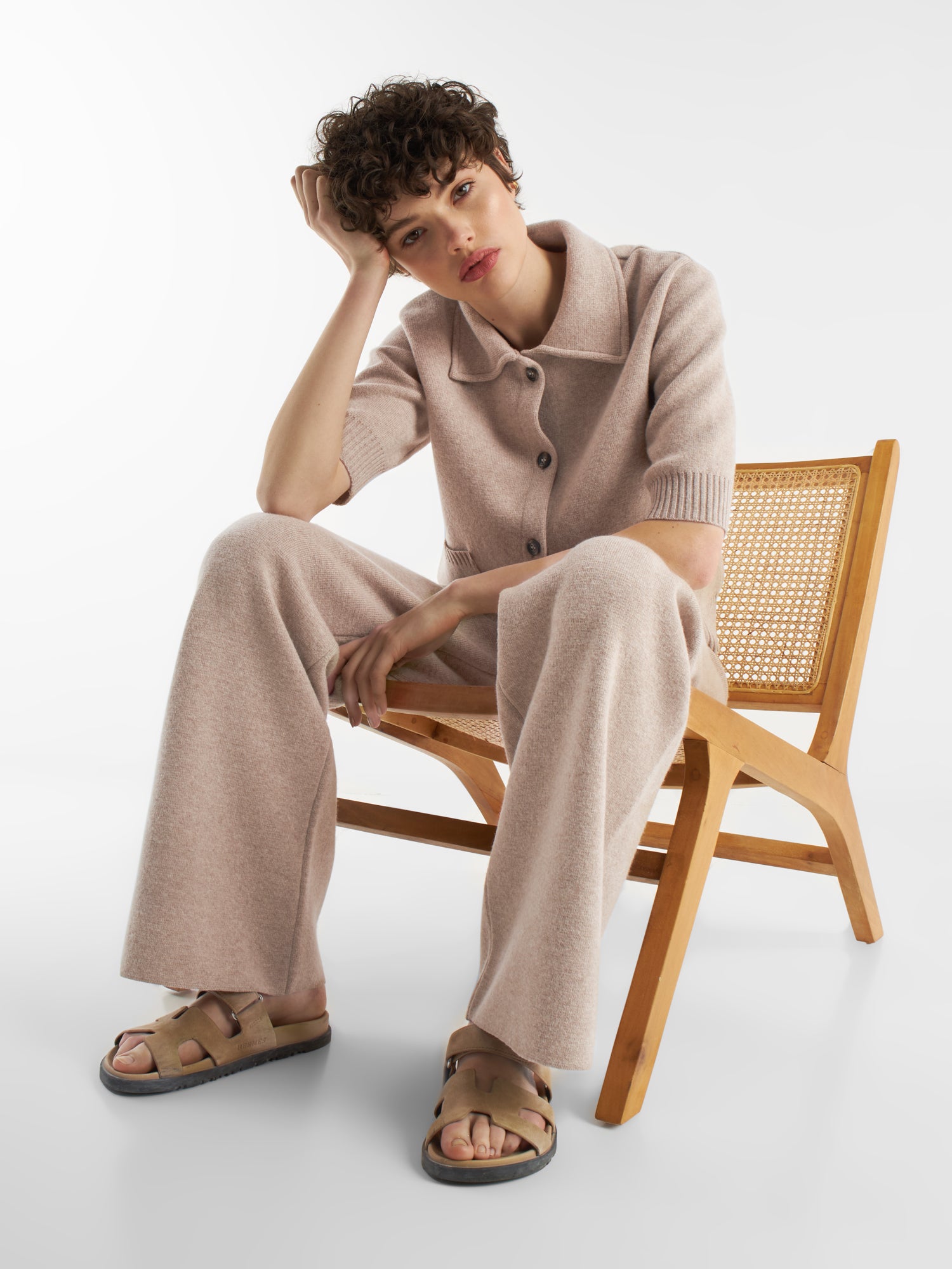 Eine Person mit kurzen lockigen Haaren sitzt in einem beigen Strickoutfit, hellbraunen Sandalen und der Jacke Lilly beige auf einem Holzstuhl, stützt den Kopf auf eine Hand und schaut nachdenklich in die Kamera.