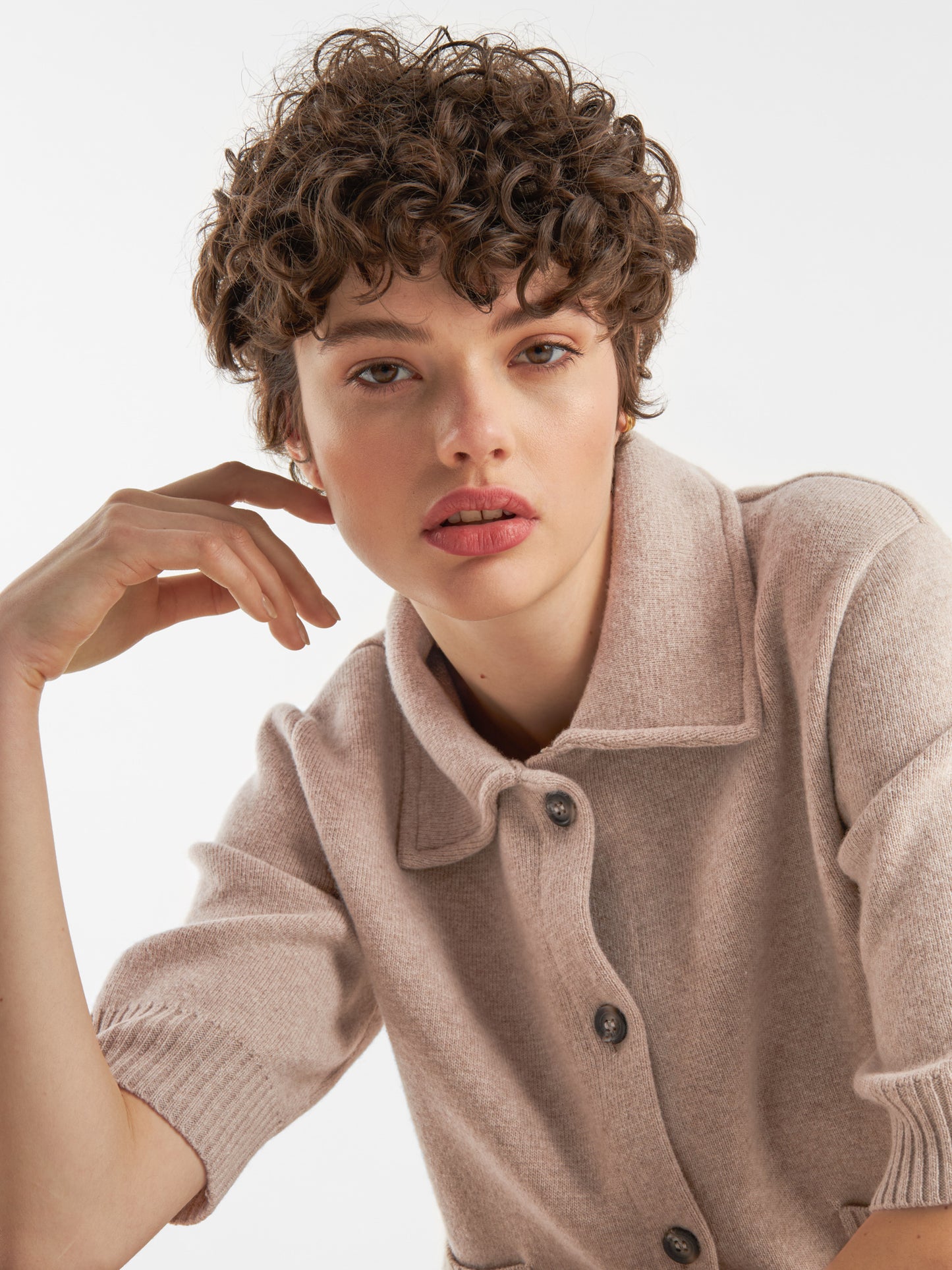 Eine Person mit kurzen, lockigen braunen Haaren trägt die Jacke Lilly beige über einem hellen Strickoberteil. Sie posiert mit einer Hand in der Nähe ihres Gesichts und schaut direkt in die Kamera vor einem schlichten weißen Hintergrund.