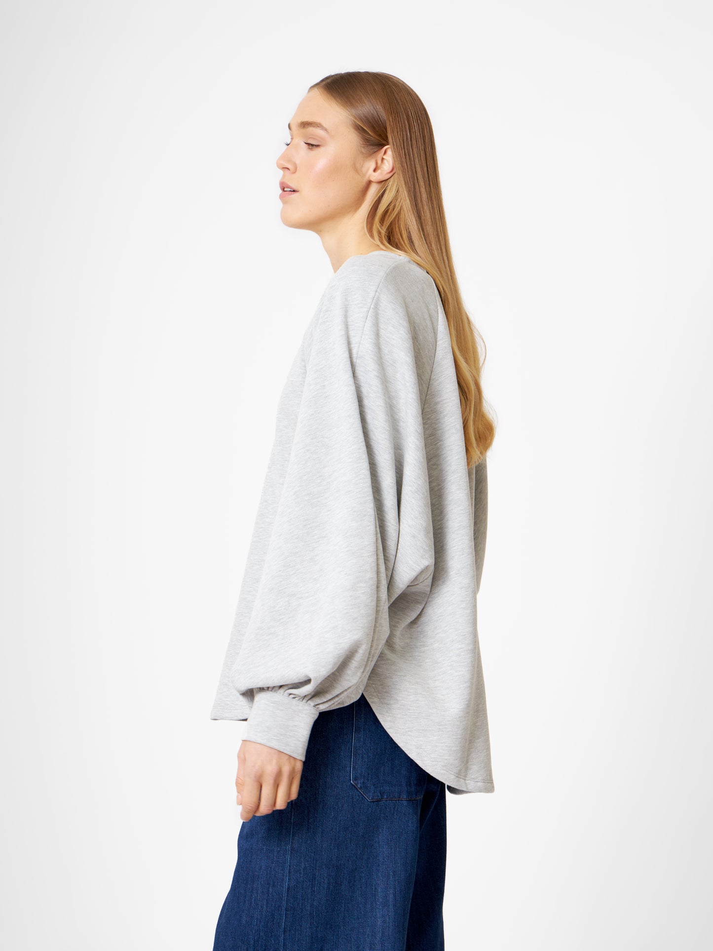 Eine Frau mit langen blonden Haaren, die das Sweatshirt Antonia Grey Mélange und Blue Jeans trägt, steht im Profil vor einem schlichten weißen Hintergrund.