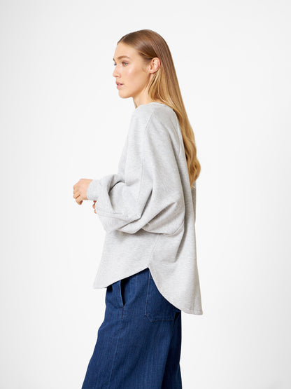 Eine Frau mit langem, hellbraunem Haar trägt ein locker sitzendes Sweatshirt Antonia Grey Mélange und eine blaue Hose mit weitem Bein und steht seitlich vor einem schlichten weißen Hintergrund.