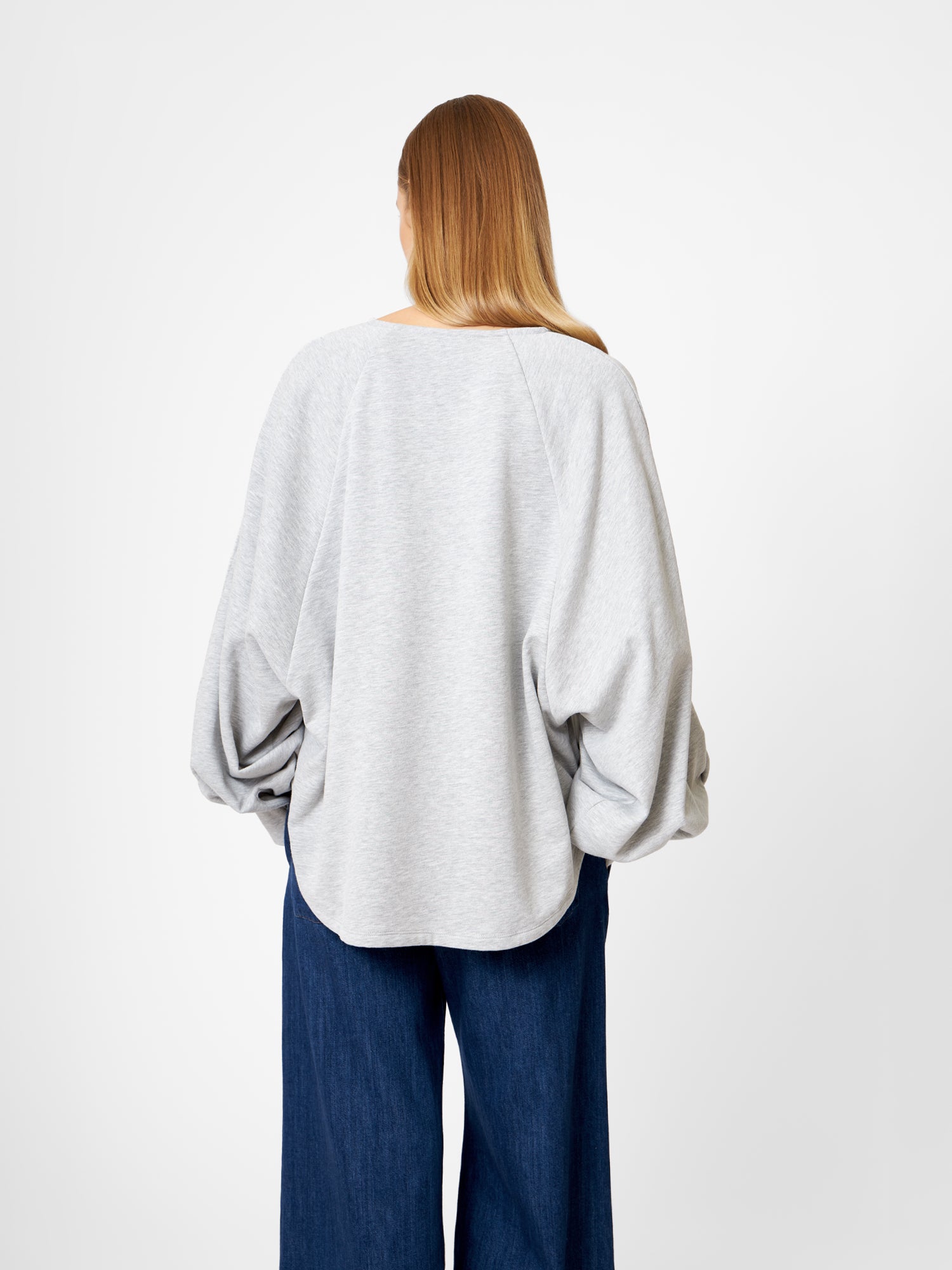 Eine Person mit langen, glatten Haaren, von hinten gesehen, trägt das Sweatshirt Antonia Grey Mélange mit weiten Ärmeln und eine dunkelblaue Hose mit weitem Bein vor einem schlichten weißen Hintergrund.