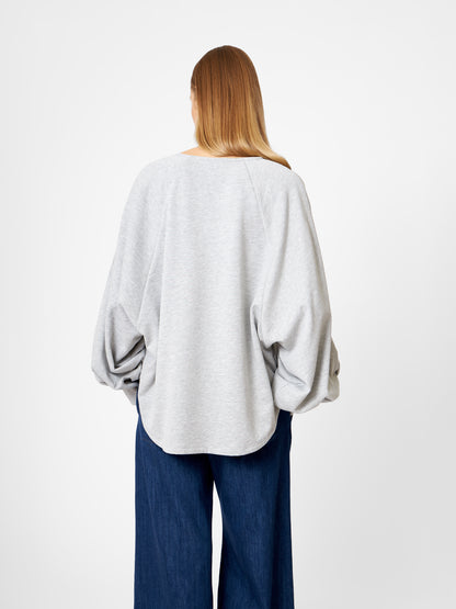 Eine Person mit langen, glatten Haaren, von hinten gesehen, trägt das Sweatshirt Antonia Grey Mélange mit weiten Ärmeln und eine dunkelblaue Hose mit weitem Bein vor einem schlichten weißen Hintergrund.