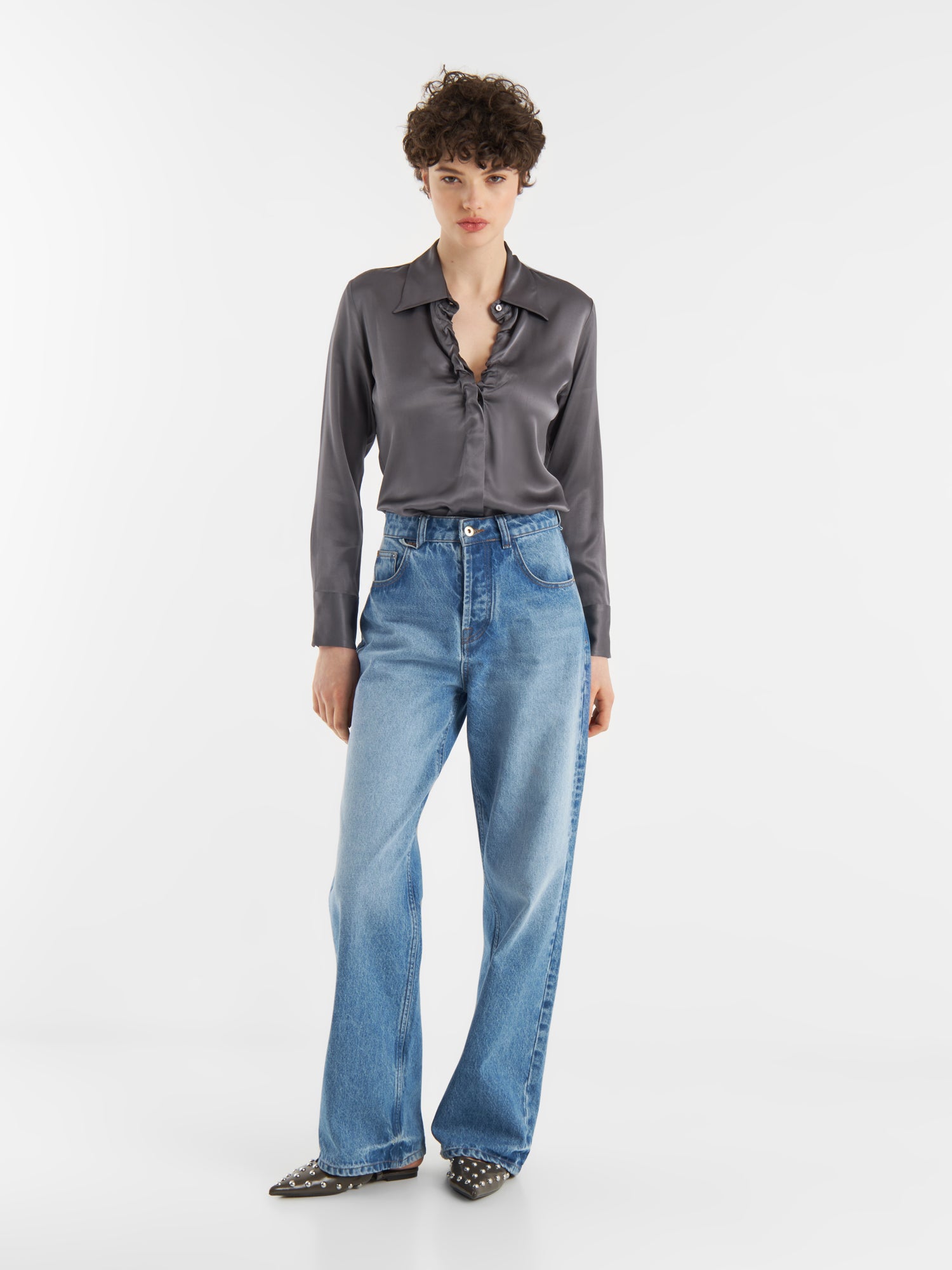 Eine Person mit kurzen lockigen Haaren trägt die Bluse Ella Shiny Grey mit einem Tunika-Ausschnitt und hellblauen, hoch sitzenden Jeans mit weitem Bein. Sie vervollständigt den Look mit spitzen schwarzen Schuhen mit silbernen Nieten, die auf einem schlichten weißen Hintergrund stehen.