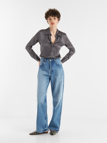 Eine Person mit kurzen lockigen Haaren trägt die Bluse Ella Shiny Grey, weite Bluejeans und schwarze spitze Stollenschuhe und steht mit den Händen in den Hüften vor einem schlichten weißen Hintergrund.