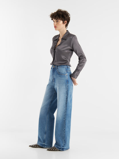 Eine Person mit kurzen lockigen Haaren trägt die Bluse Ella Shiny Grey mit einem Tunika-Ausschnitt, die sie in eine hoch taillierte, weit geschnittene Bluejeans und schwarze Flats mit kleinen weißen Punkten steckt, und steht vor einem schlichten weißen Hintergrund.