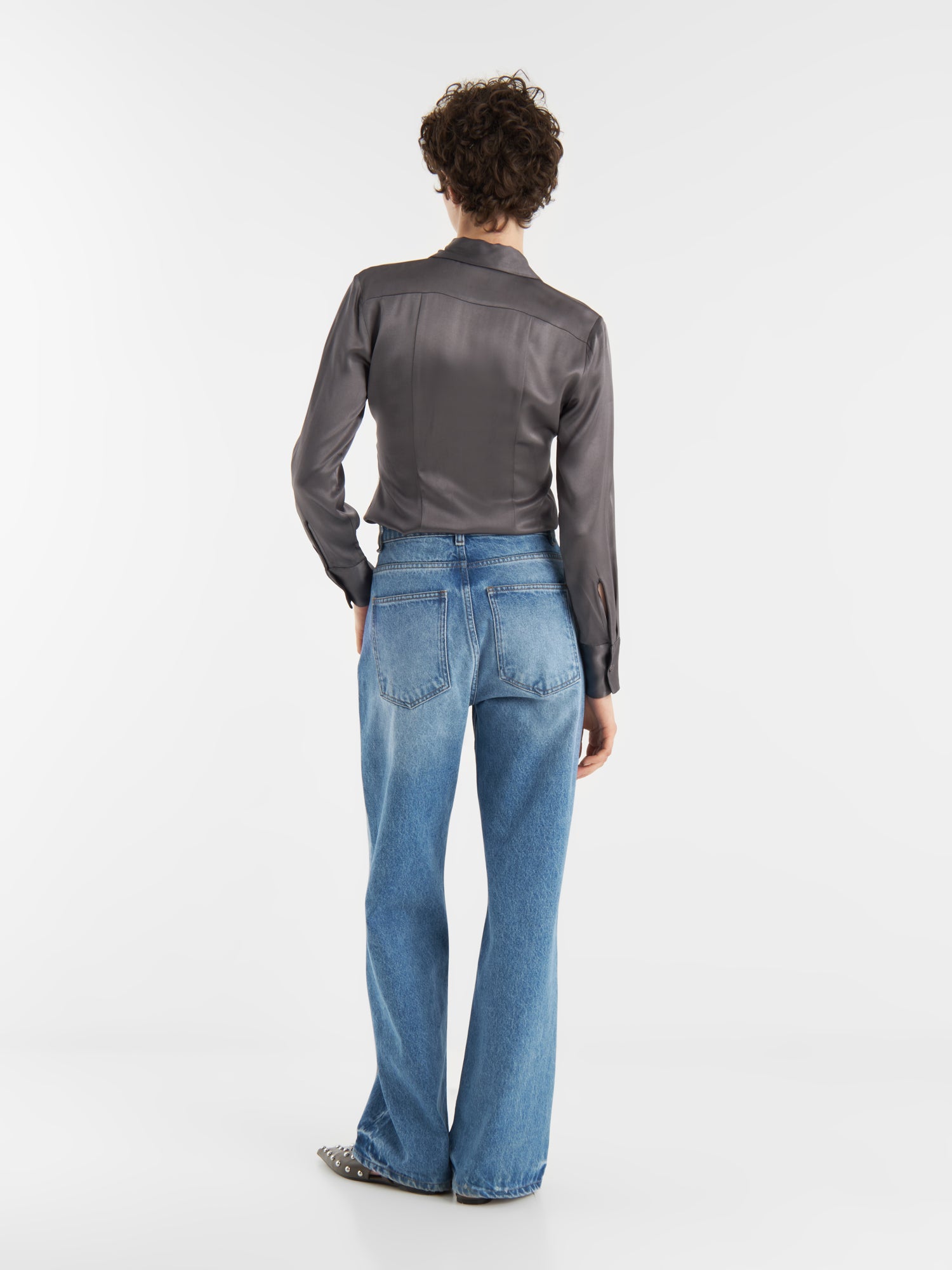 Eine Person mit kurzen lockigen Haaren, die die Bluse Ella Shiny Grey mit Tunika-Ausschnitt und hochgezogenen Bluejeans trägt, steht mit dem Gesicht nach unten auf einem einfarbig weißen Hintergrund.