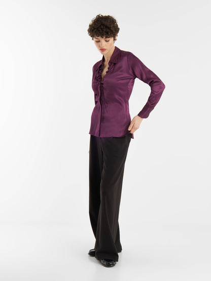 Eine Person mit kurzen lockigen Haaren trägt die Bluse Ella Shiny Purple mit Rüschenbesatz, eine schwarze Hose mit weitem Bein und schwarze Schuhe und steht vor einem schlichten weißen Hintergrund.