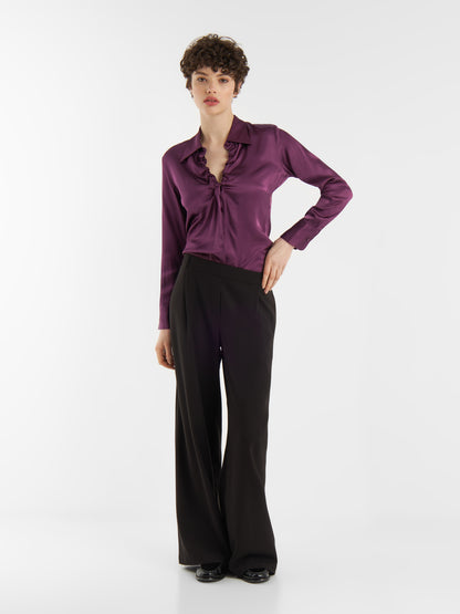 Eine Frau mit kurzen lockigen Haaren trägt die Bluse Ella Shiny Purple, eine langärmelige Satinbluse mit Rüschenkragen, gepaart mit einer schwarzen Hose mit hoher Taille und weiten Beinen und schwarzen Schuhen, vor einem schlichten weißen Hintergrund.