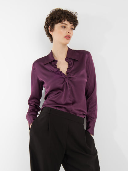 Eine Person mit kurzen lockigen Haaren trägt die Bluse Ella Shiny Purple mit einem gerüschten Ausschnitt und einer schwarzen Hose mit hoher Taille und posiert vor einem schlichten weißen Hintergrund.