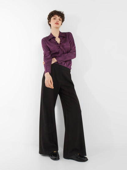 Eine Person mit kurzen lockigen Haaren trägt die Bluse Ella Shiny Purple und eine schwarze Hose mit weitem Bein. Sie steht vor einem schlichten weißen Hintergrund, hat einen Arm gekreuzt und schaut selbstbewusst in die Kamera.