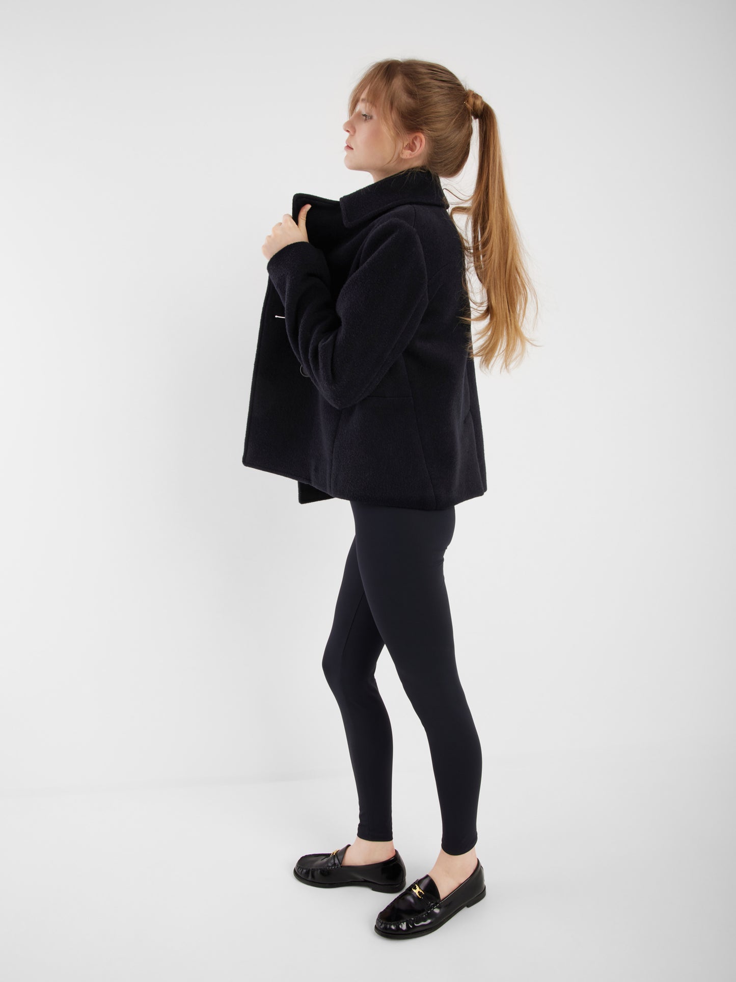 Eine Frau mit langen Haaren in einem Pferdeschwanz steht im Profil und trägt die Jacke Caban Navy, schwarze Leggings und schwarze Slipper vor einem schlichten weißen Hintergrund.