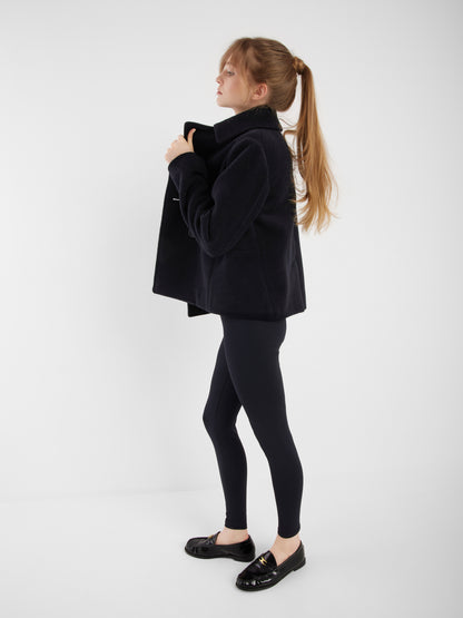 Eine Frau mit langen Haaren in einem Pferdeschwanz steht im Profil und trägt die Jacke Caban Navy, schwarze Leggings und schwarze Slipper vor einem schlichten weißen Hintergrund.