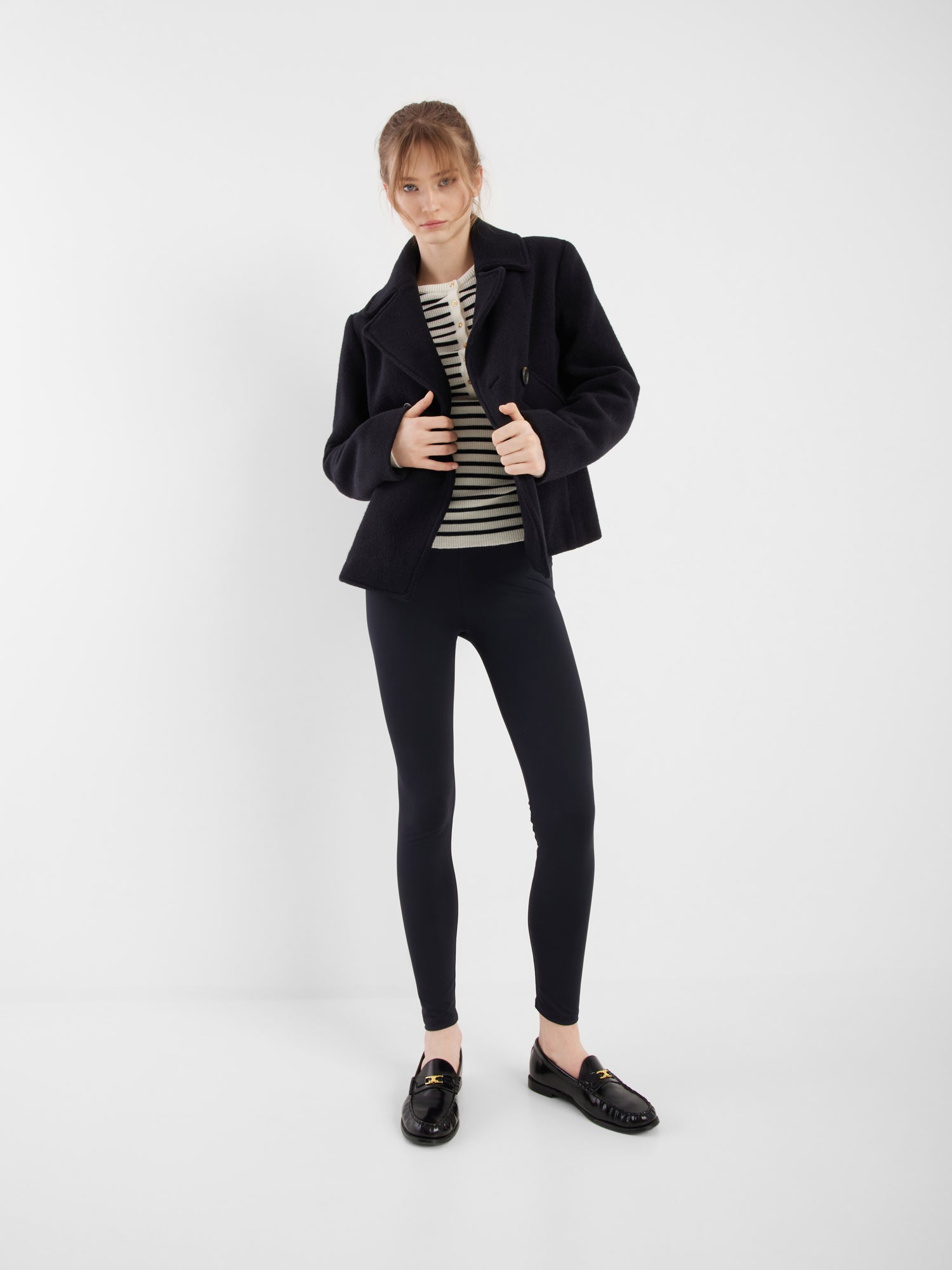 Eine Frau steht vor einem weißen Hintergrund und trägt die Jacke Caban Navy, gepaart mit einem schwarz-weiß gestreiften Oberteil, schwarzen Leggings und schwarzen Halbschuhen. Sie blickt nach vorne und rückt ihre Jacke zurecht.
