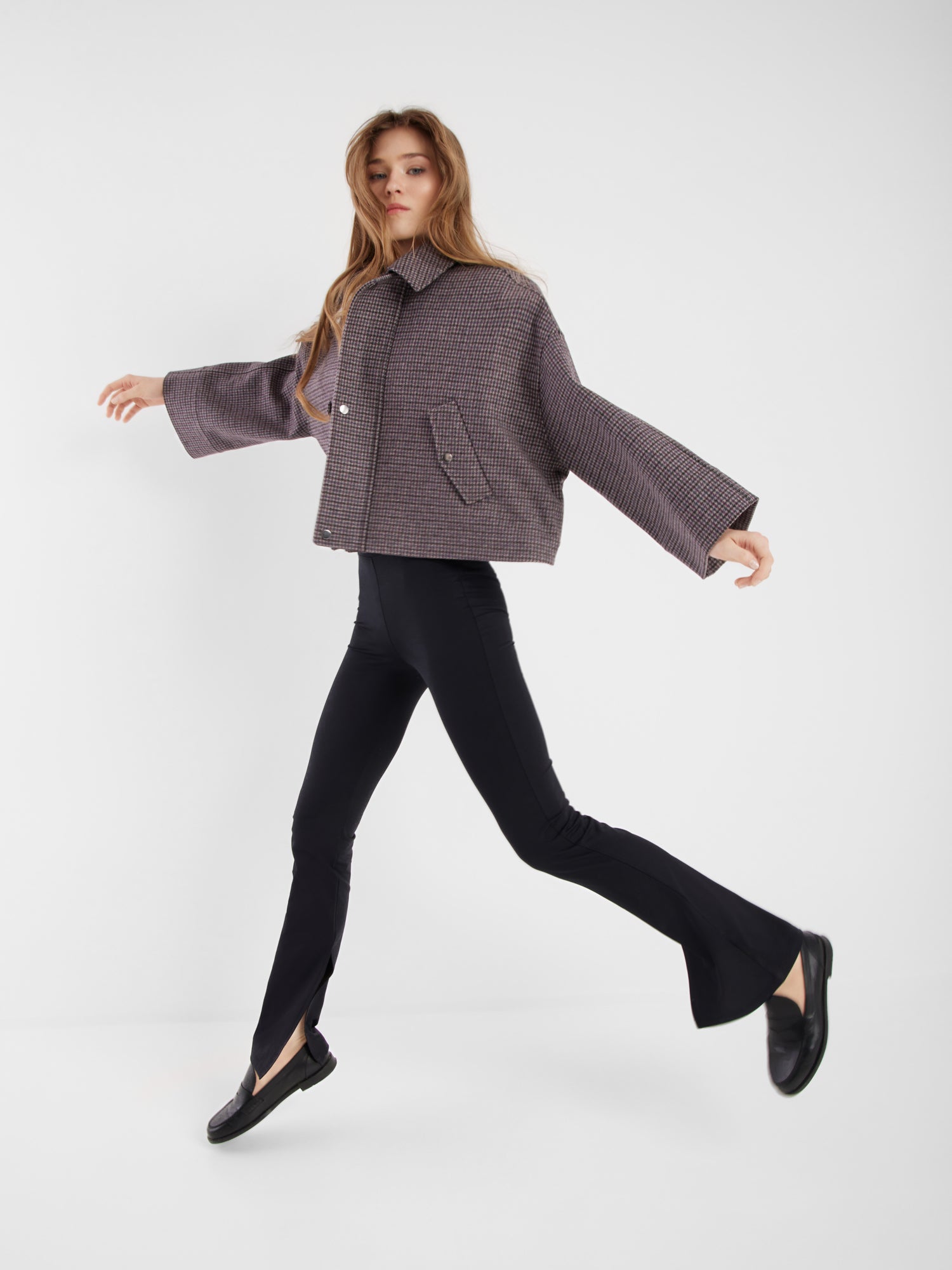 Eine Frau mit langen Haaren geht energisch spazieren. Sie trägt die Jacke Victoria mit Hahnentrittmuster, schwarze Schlaghosen und schwarze Slipper vor einem schlichten weißen Hintergrund.